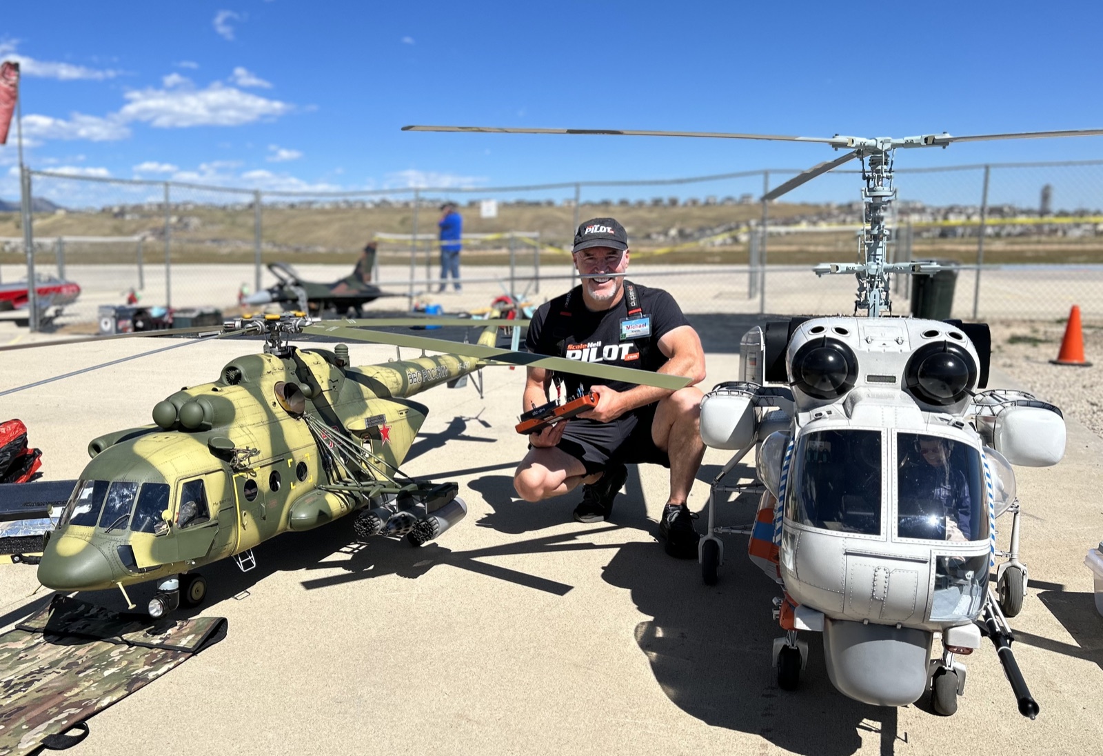 Michael Kranitz at a scale RC helicopter flying field, crouched with his transmitter between a camouflaged Mi-8 and a silver Ka-27 scale helicopter